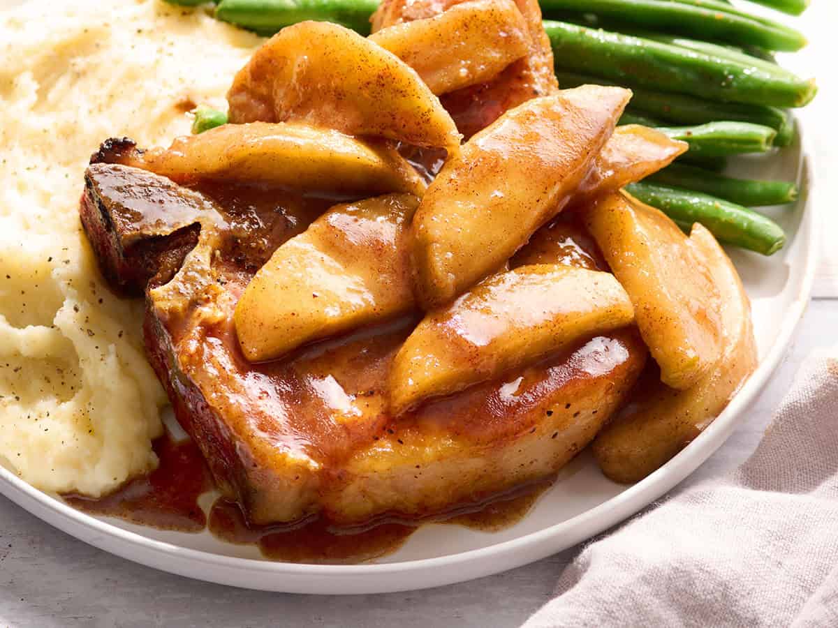 Side view of a pork chop, topped with fried apples, on a plate with mashed potatoes and green beans.