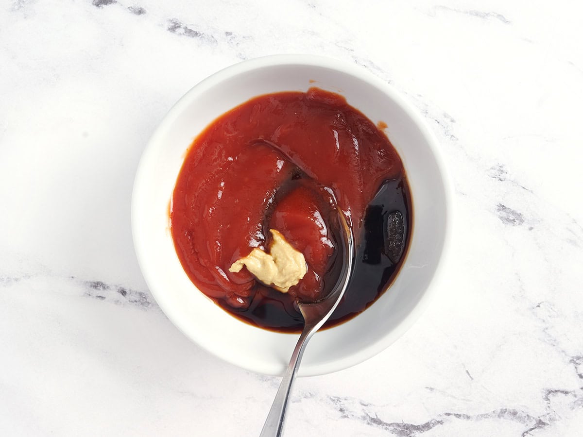Dijon mustard, ketchup, soy sauce, and brown sugar being mixed together with a spoon in a bowl.