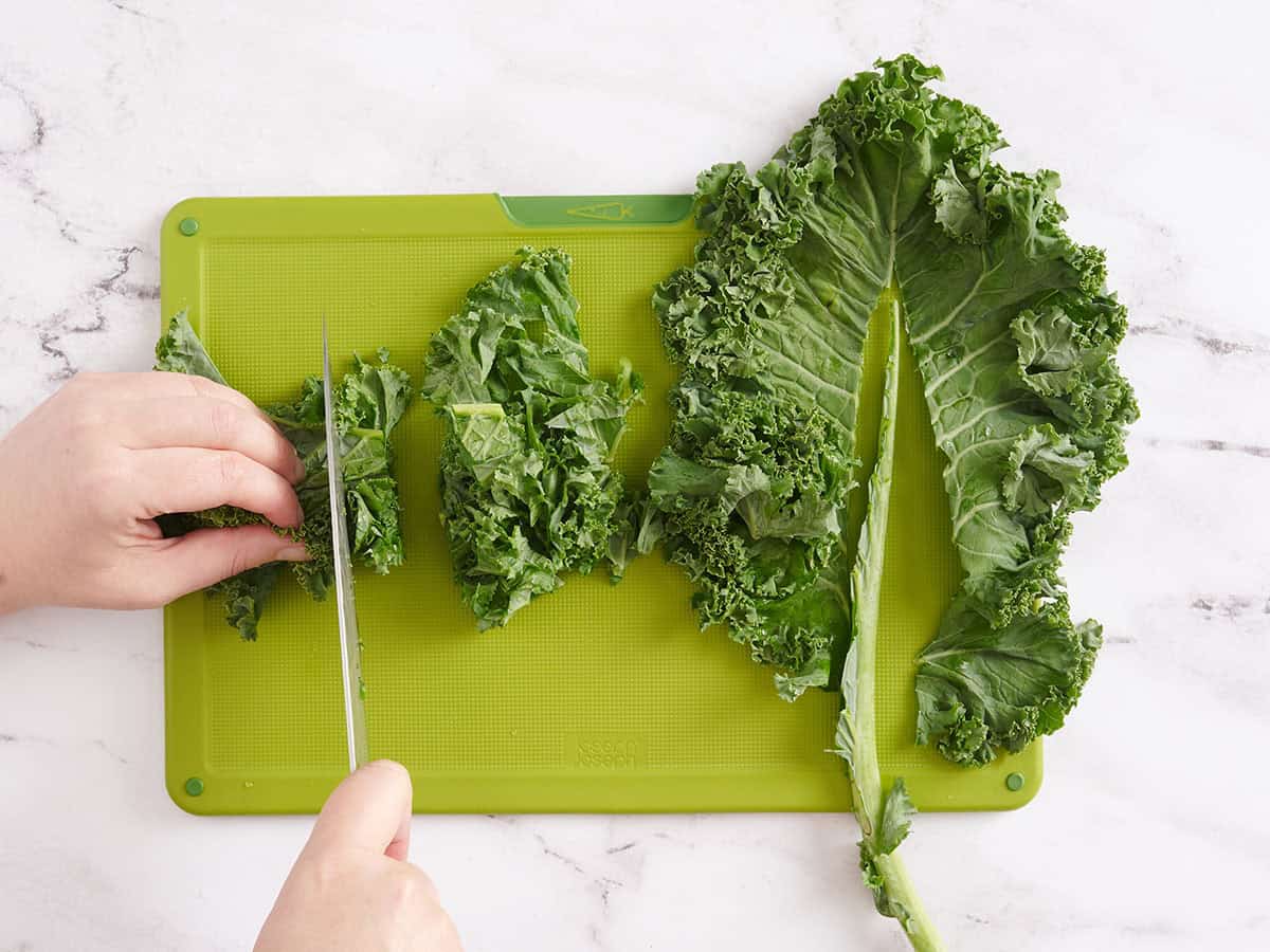A hand using a knife to slice fresh kale on a green cutting board.