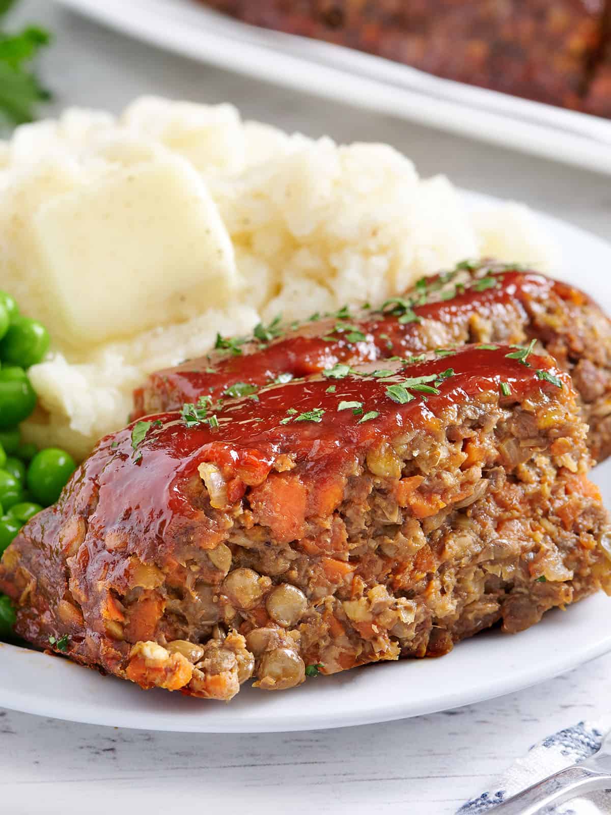 A slice of lentil loaf on a plate with peas and mashed potato.