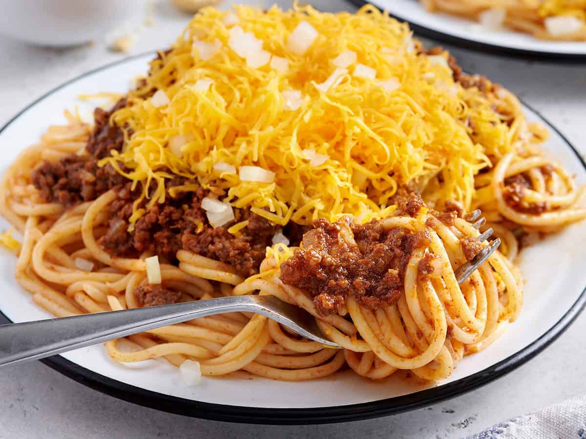 Side view of a plate of homemade skyline chili with shredded cheese, diced onions, and a fork taking some.