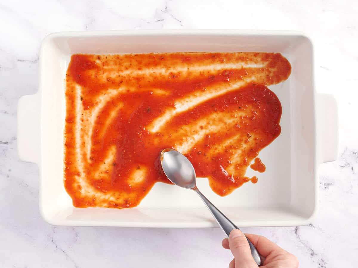A spoon spreading marinara sauce in the bottom of a casserole dish.