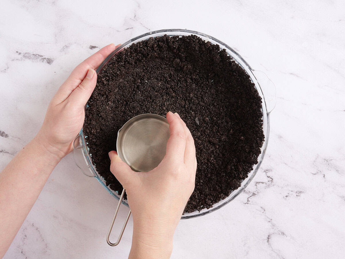 Hands using a measuring cup to press crushed cookies into a pie dish.