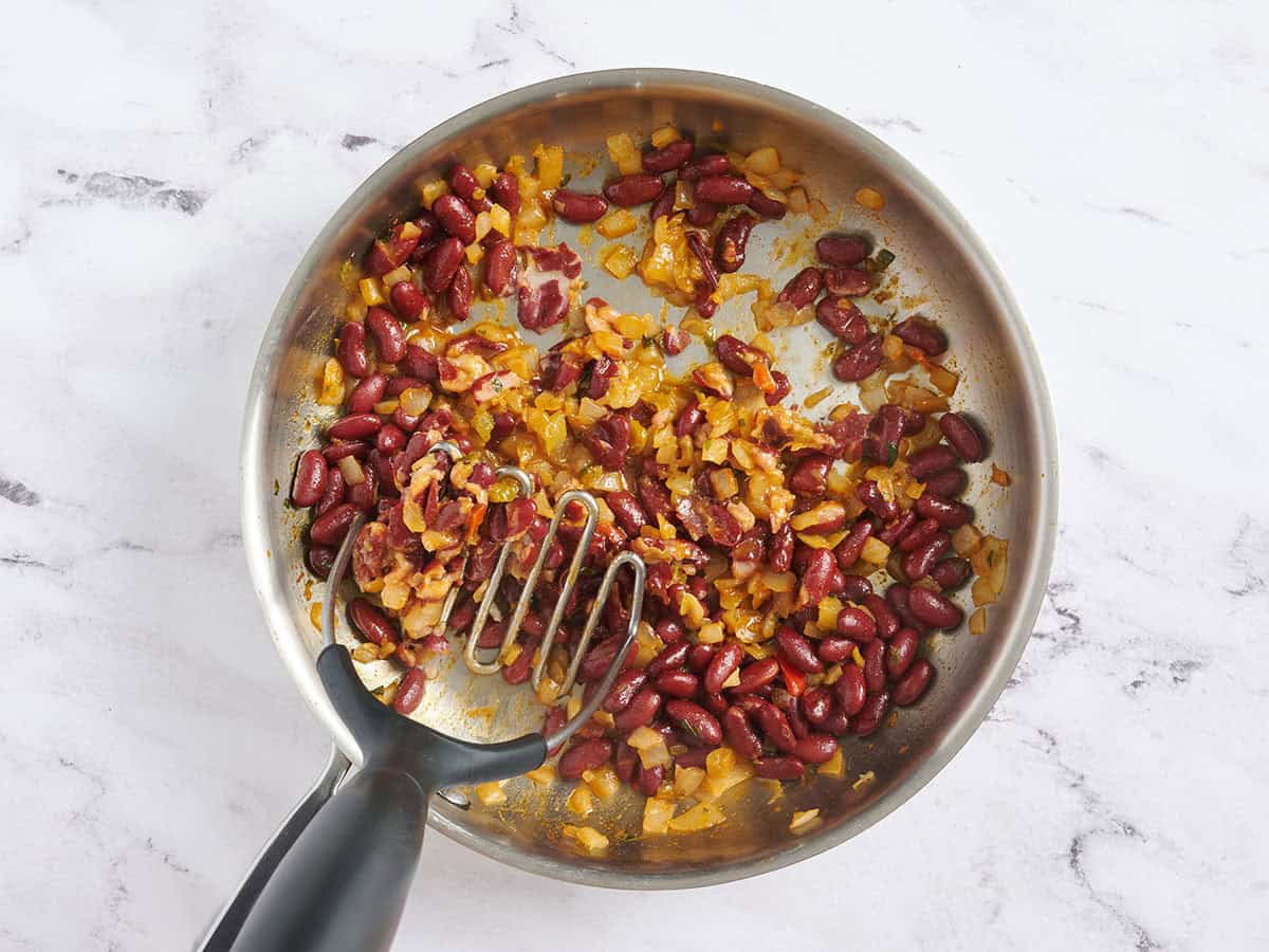 A potato masher mashing kidney beans in a skillet.