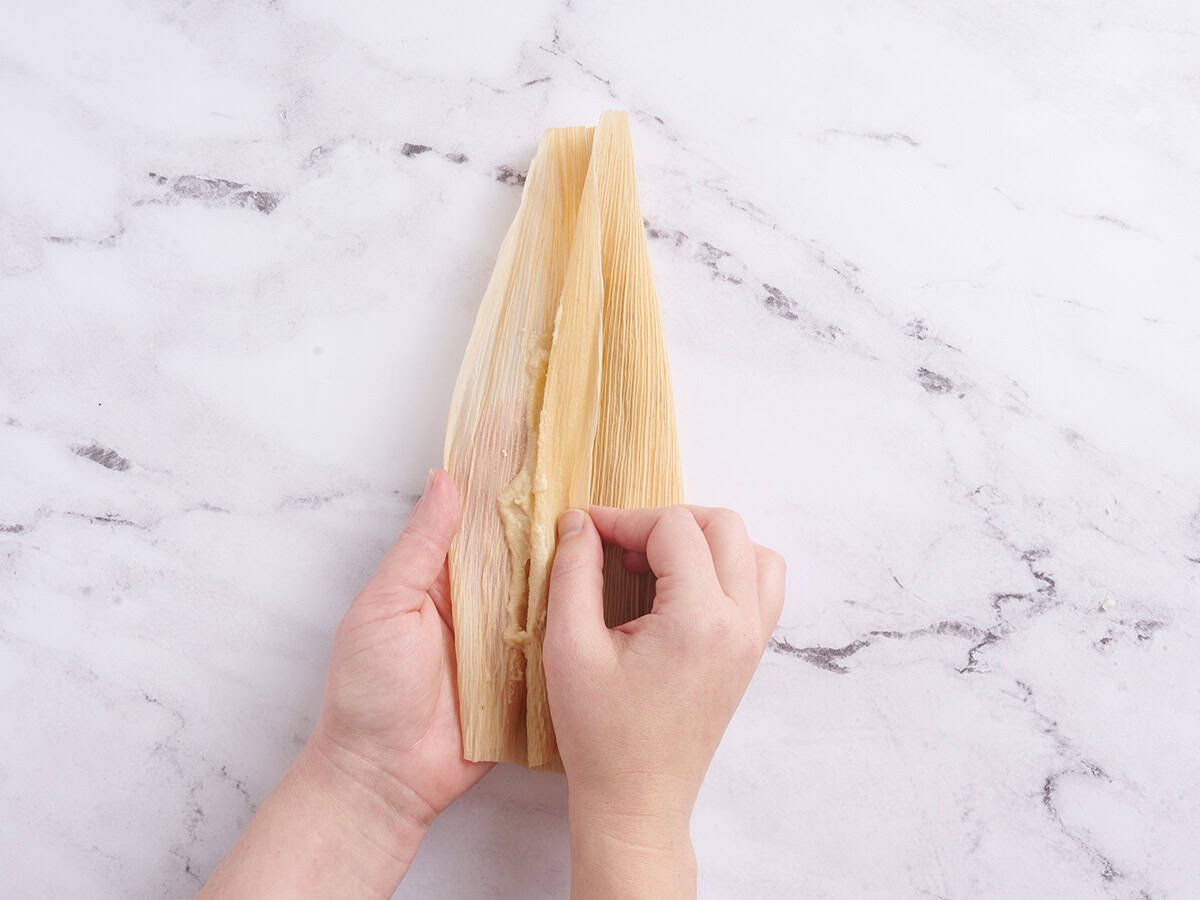 Hands folding the side of a corn husk over filling.