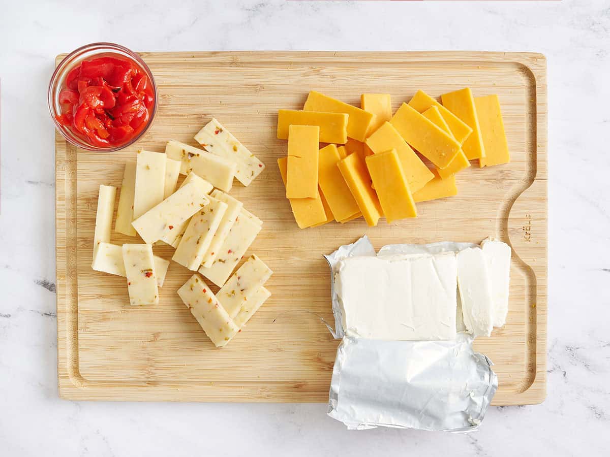 Sliced cheddar, pepper jack, and cream cheese on a wooden cutting board with a jar of red peppers.