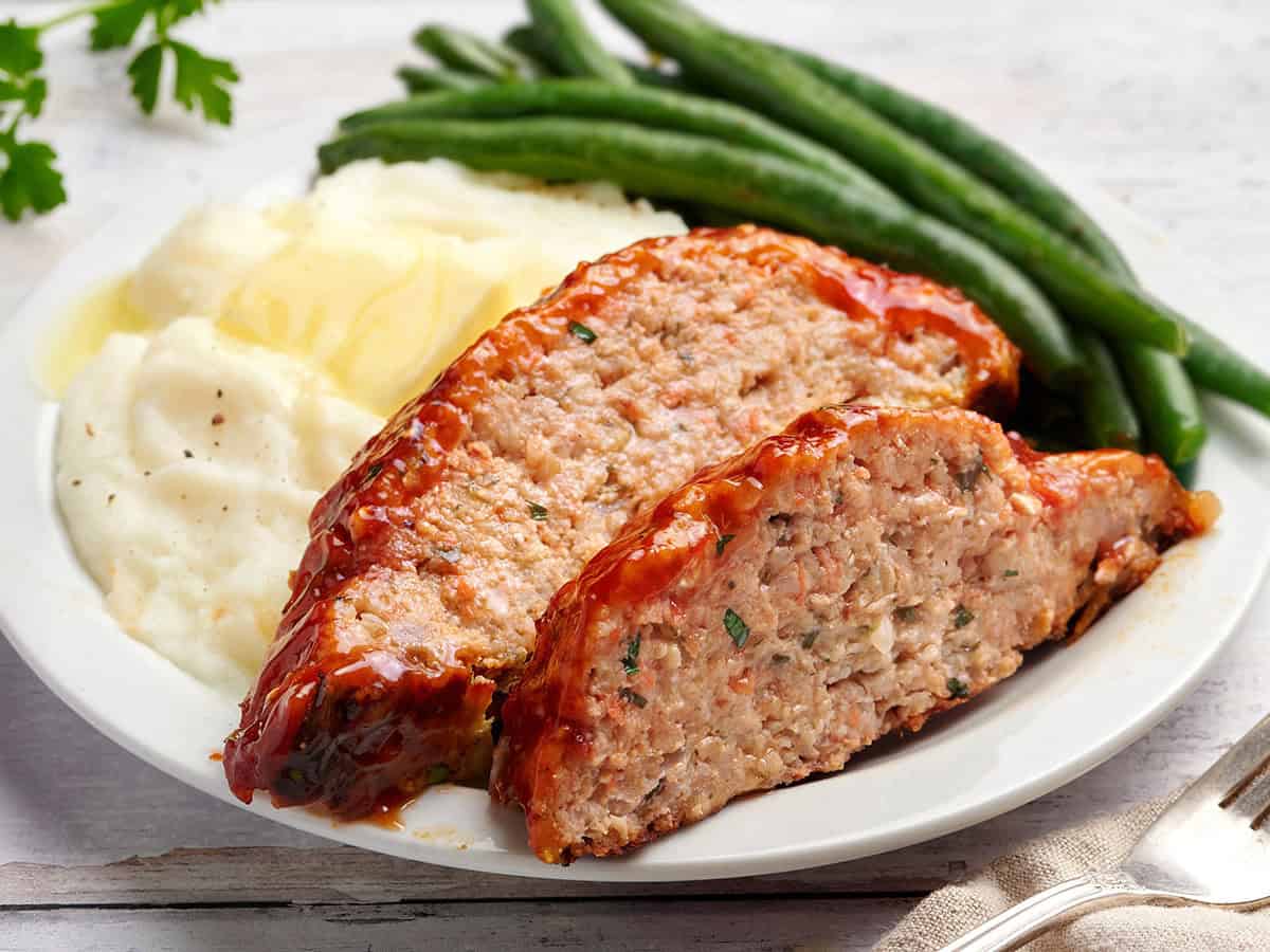 Slices of chicken meatloaf on a white plate with mashed potatoes and green beans. 