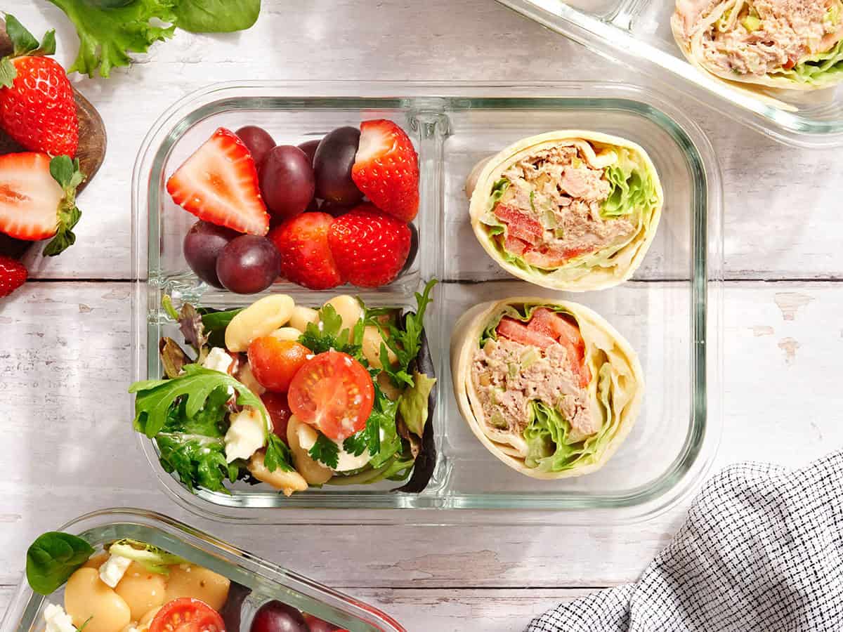 Overhead view of tuna wrap in a meal prep box with strawberries, grapes and white bean salad.