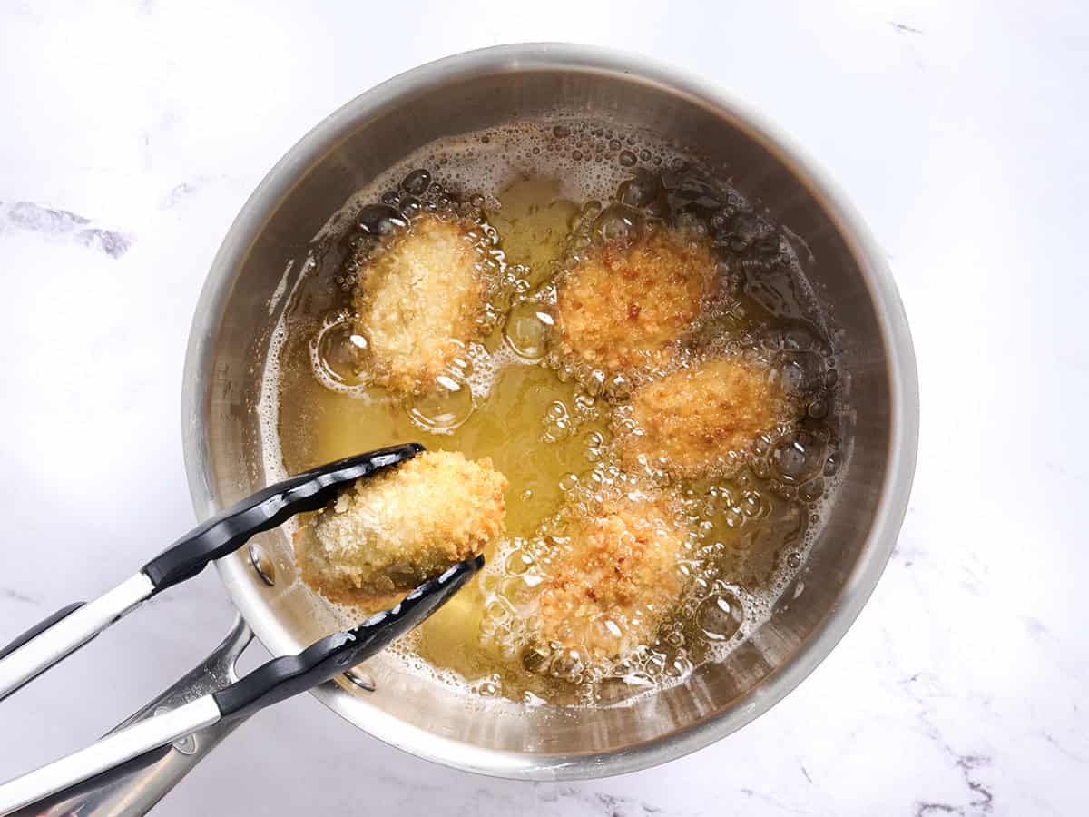 Chicken croquettes frying in oil with a tong flipping one over.