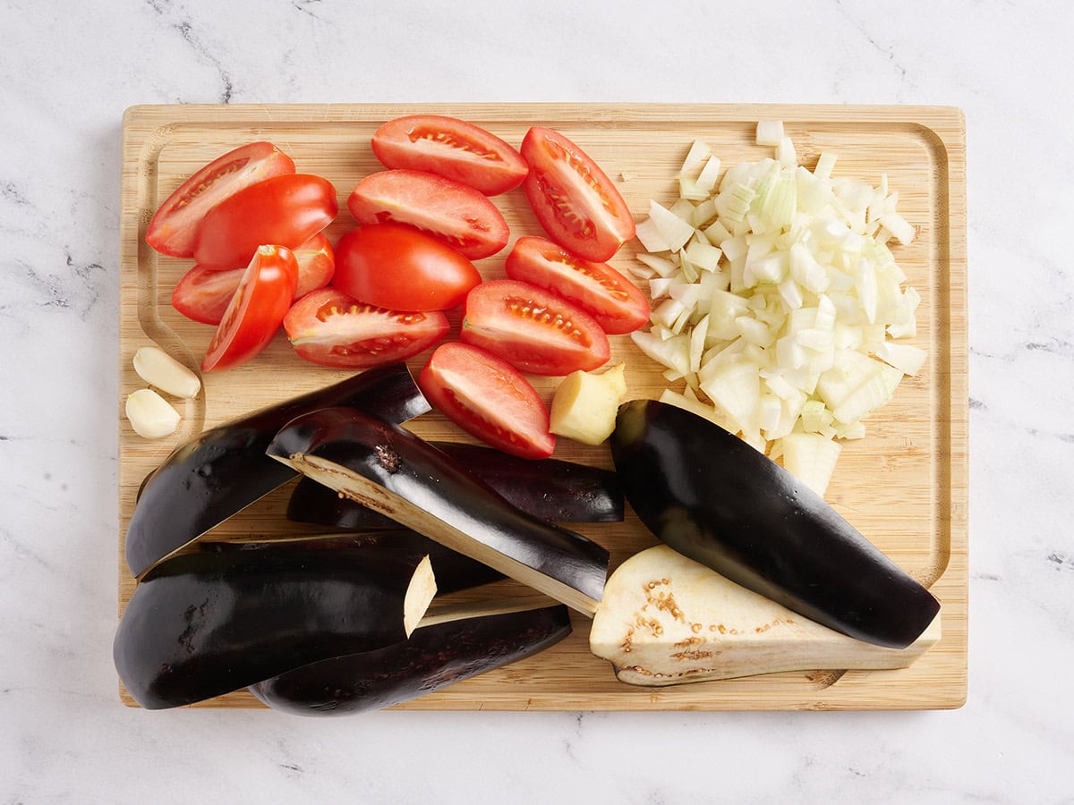 Quartered tomatoes and eggplant, diced onion and peeled garlic cloves on a wooden cutting board.