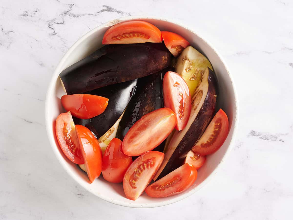 Eggplant and tomato quarters in a bowl with seasonings and oil.