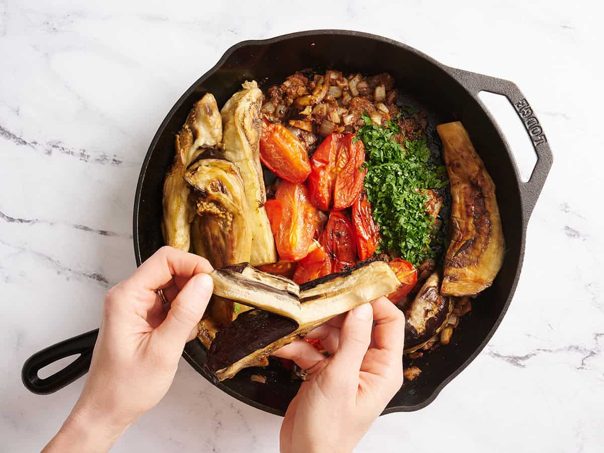 Hands peeling the skin from roasted eggplant before adding it to a skillet with tomatoes.