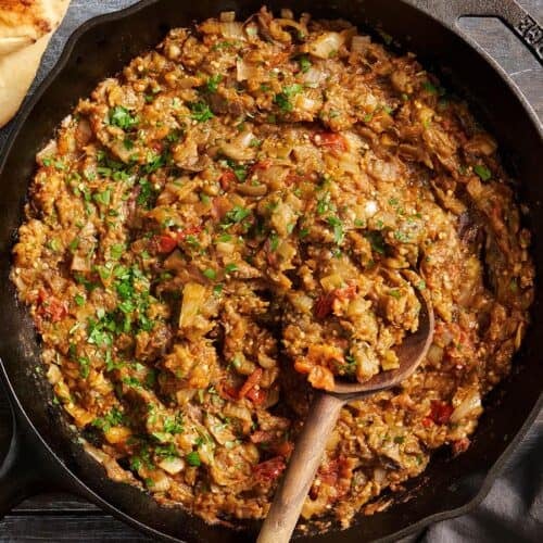 Overhead view of eggplant curry in a skillet with a wooden spoon.