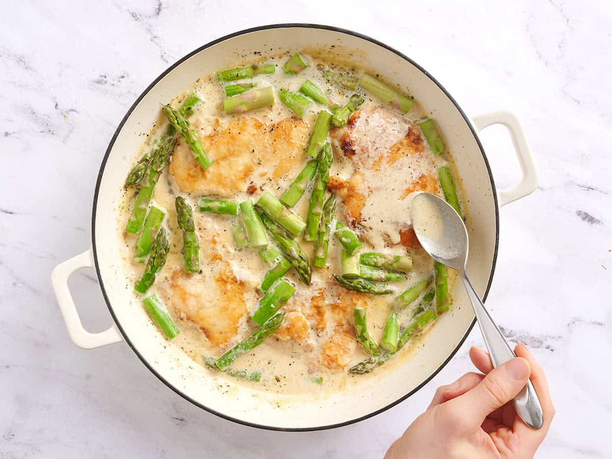 A hand spooning creamy sauce over asparagus chicken.