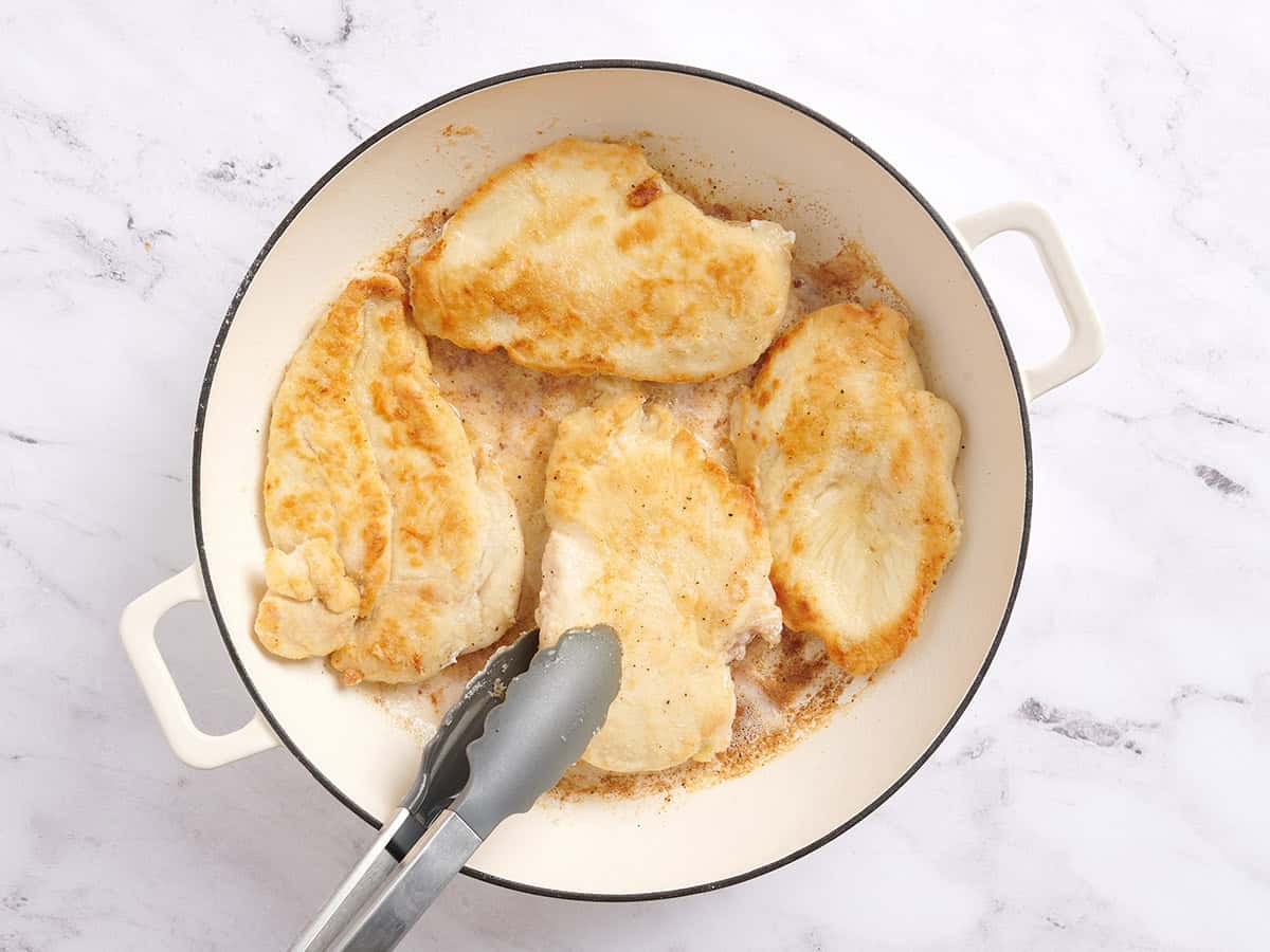Kitchen tongs flipping chicken breasts in a skillet.