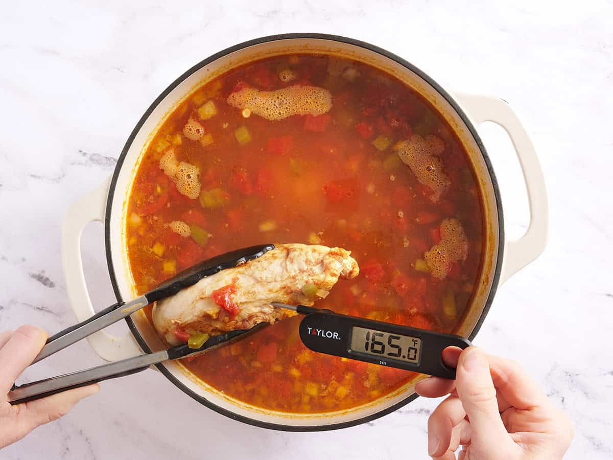 Tongs holding a cooked chicken breast over a pot of chicken taco soup and a meat thermometer checking the internal temperature of the chicken.
