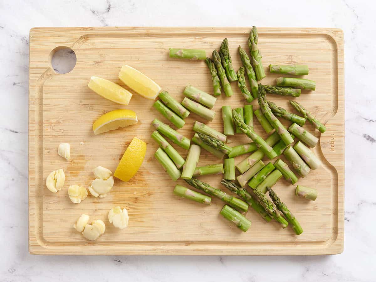 Crushed garlic cloves, sliced asparagus and sliced lemon on a wooden cutting board.