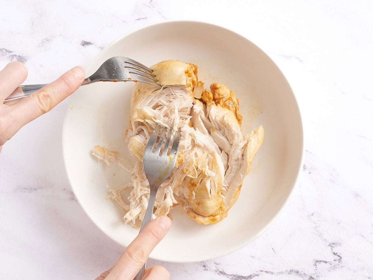 Hands using two forks to shred cooked chicken breasts in a bowl.