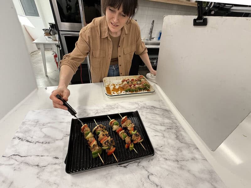 A woman checking the internal temperature of chicken kabobs on a grill with a meat thermometer.