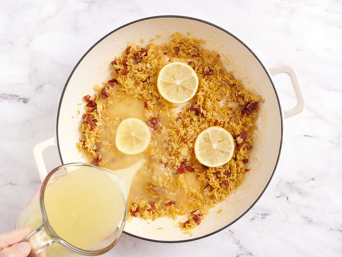 Lemon slices and onions in a skillet with chicken broth being poured in.