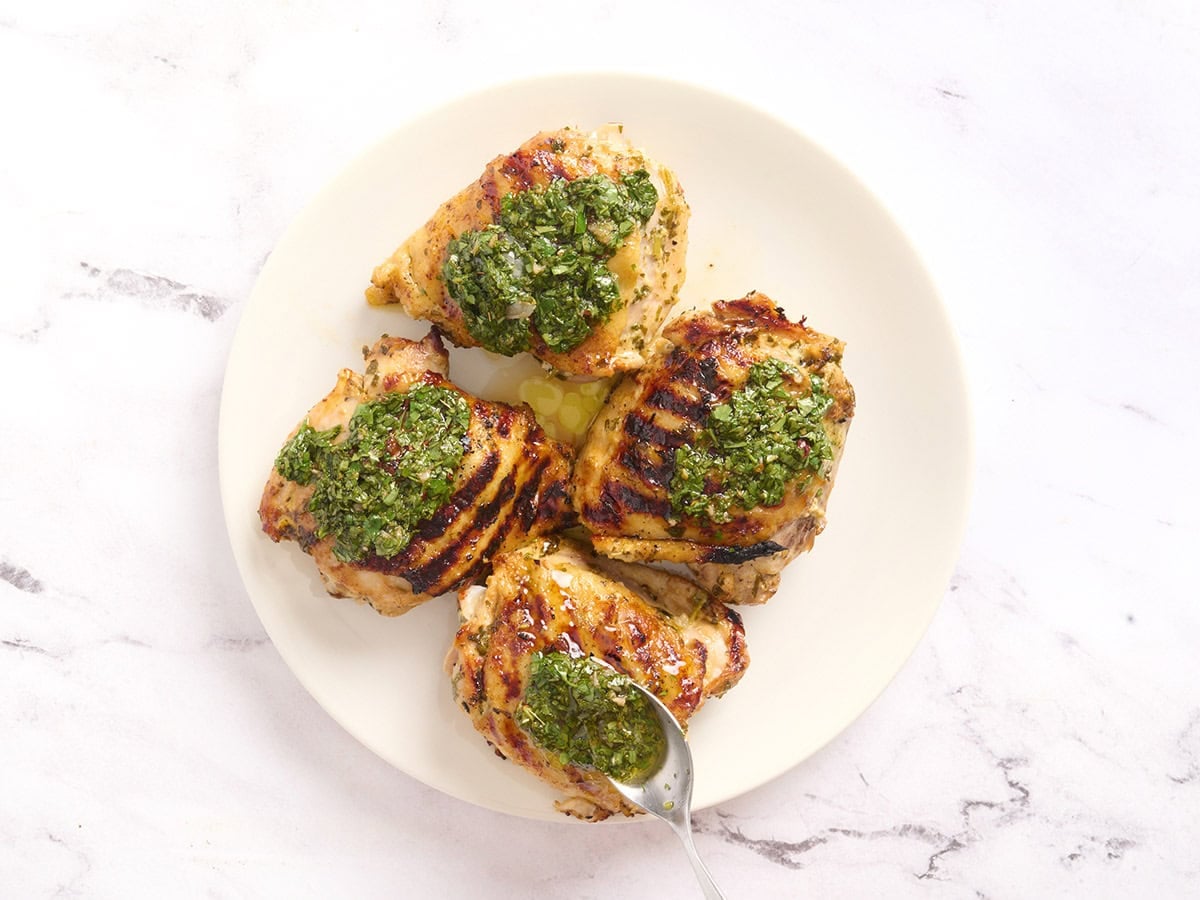 Grilled chicken thighs on a plate with a spoon adding chimichurri on top.