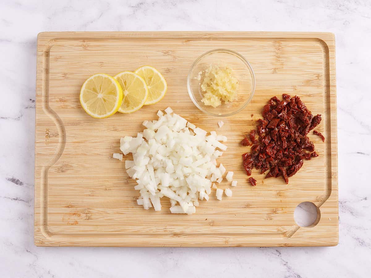 Sliced lemons, diced onion, diced sundried tomatoes, and minced garlic on a wooden cutting board.