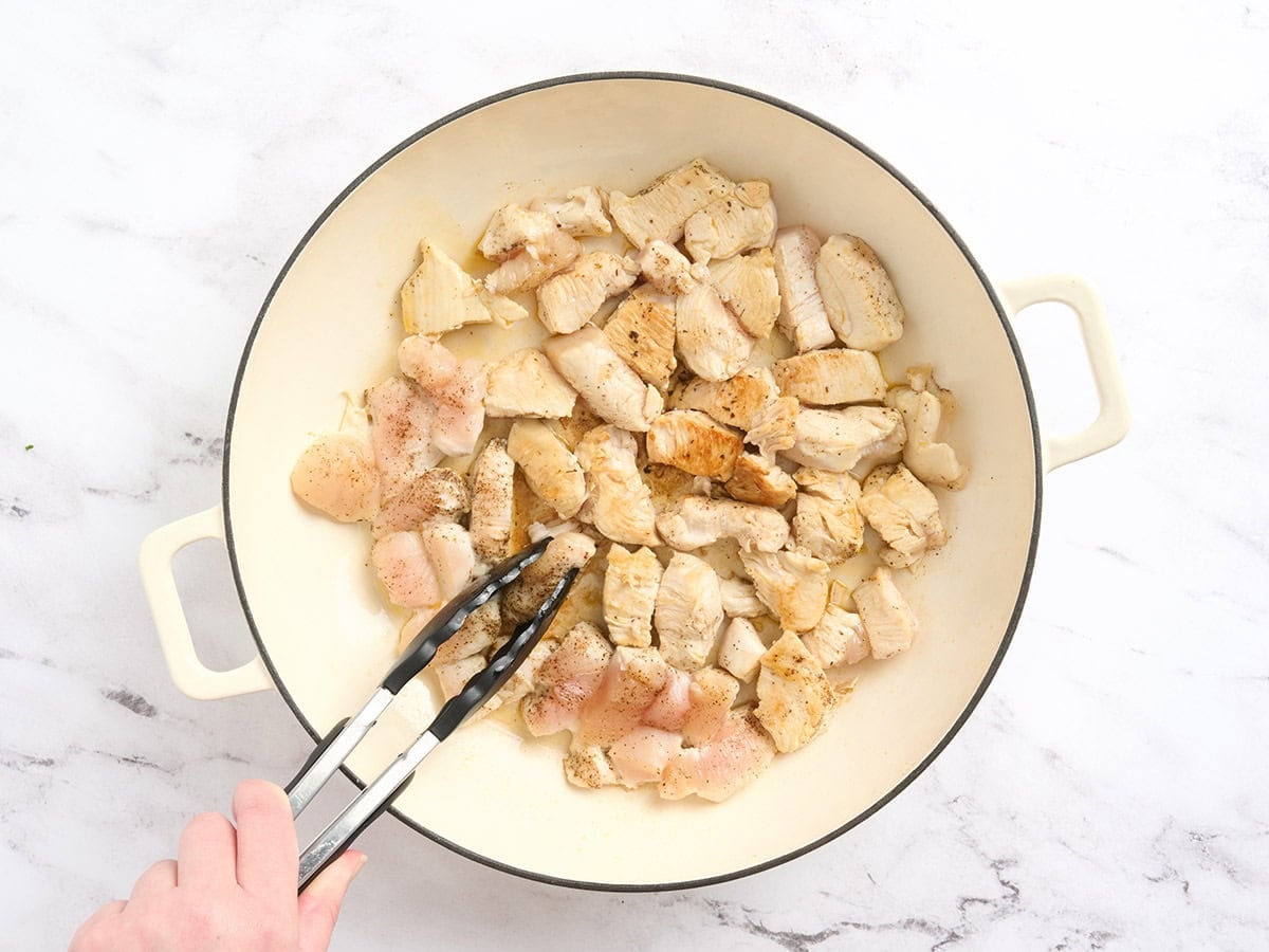 Kitchen tongs flipping pieces of chicken in a pot.
