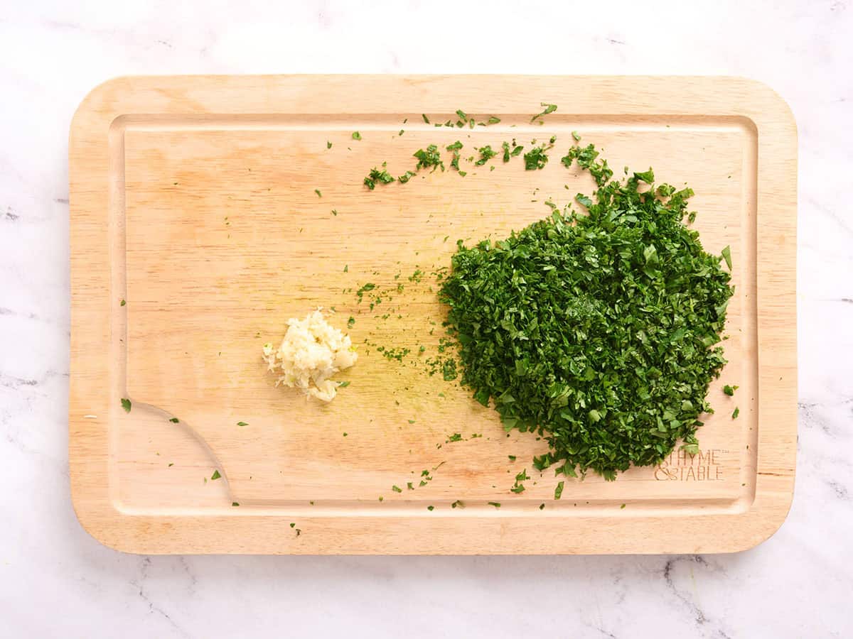 Minced herbs and garlic on a wooden cutting board.