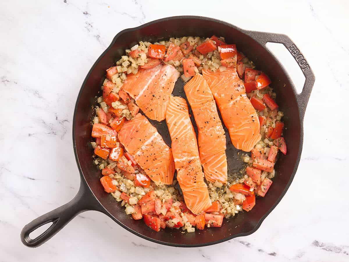 Tomatoes and onions pushed to the side and salmon fillets added to the center of a skillet.