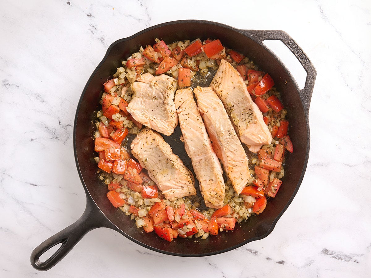 Tomatoes and onions pushed to the side and seared salmon fillets in the center of a skillet.