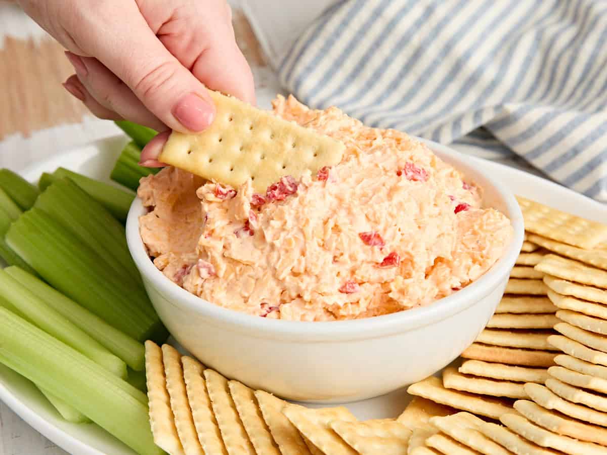 Side view of a bowl of southern pimento cheese dip with a hand dipping a cracker in.