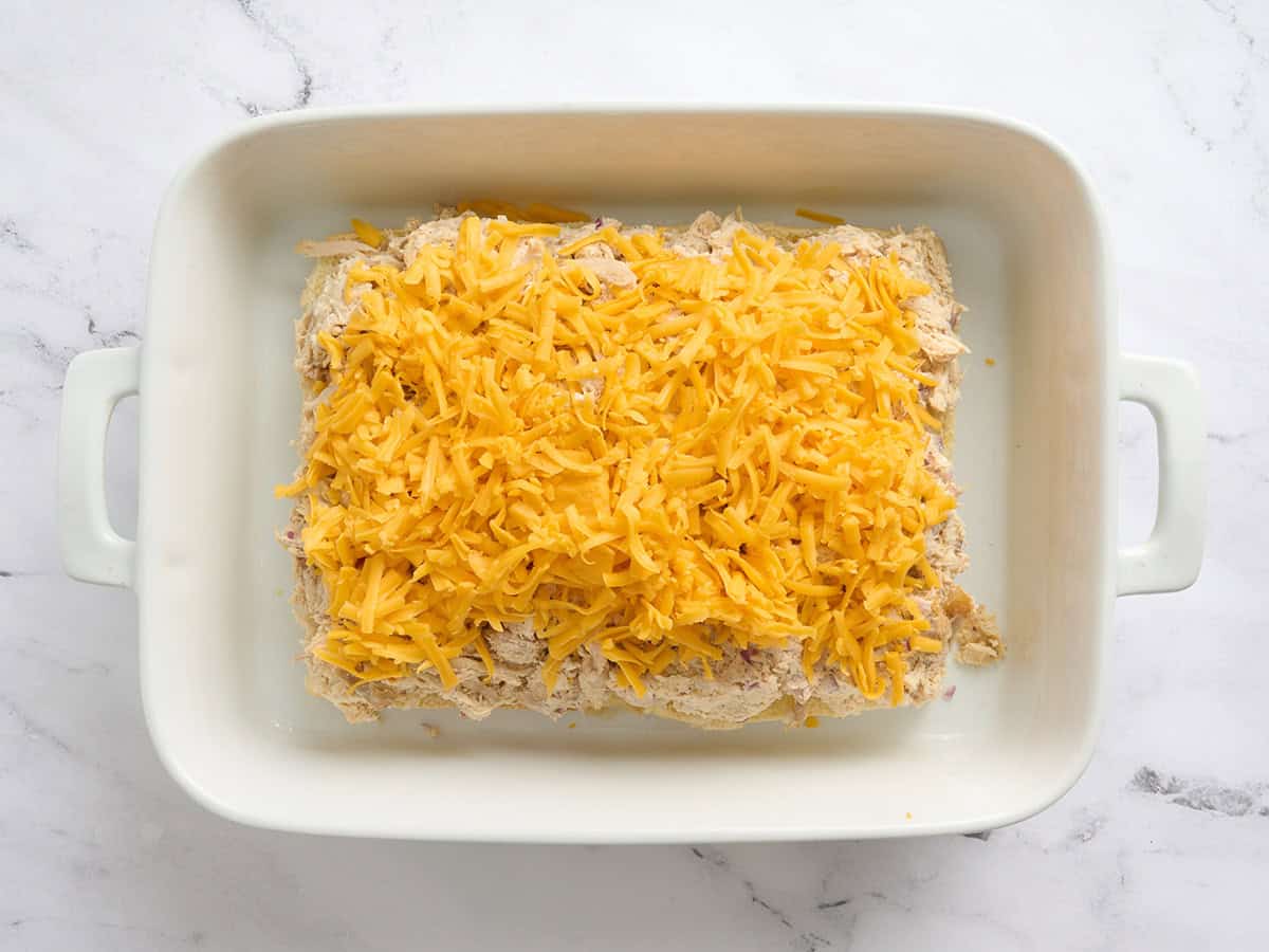 Shredded cheddar added to chicken sliders in a baking dish.