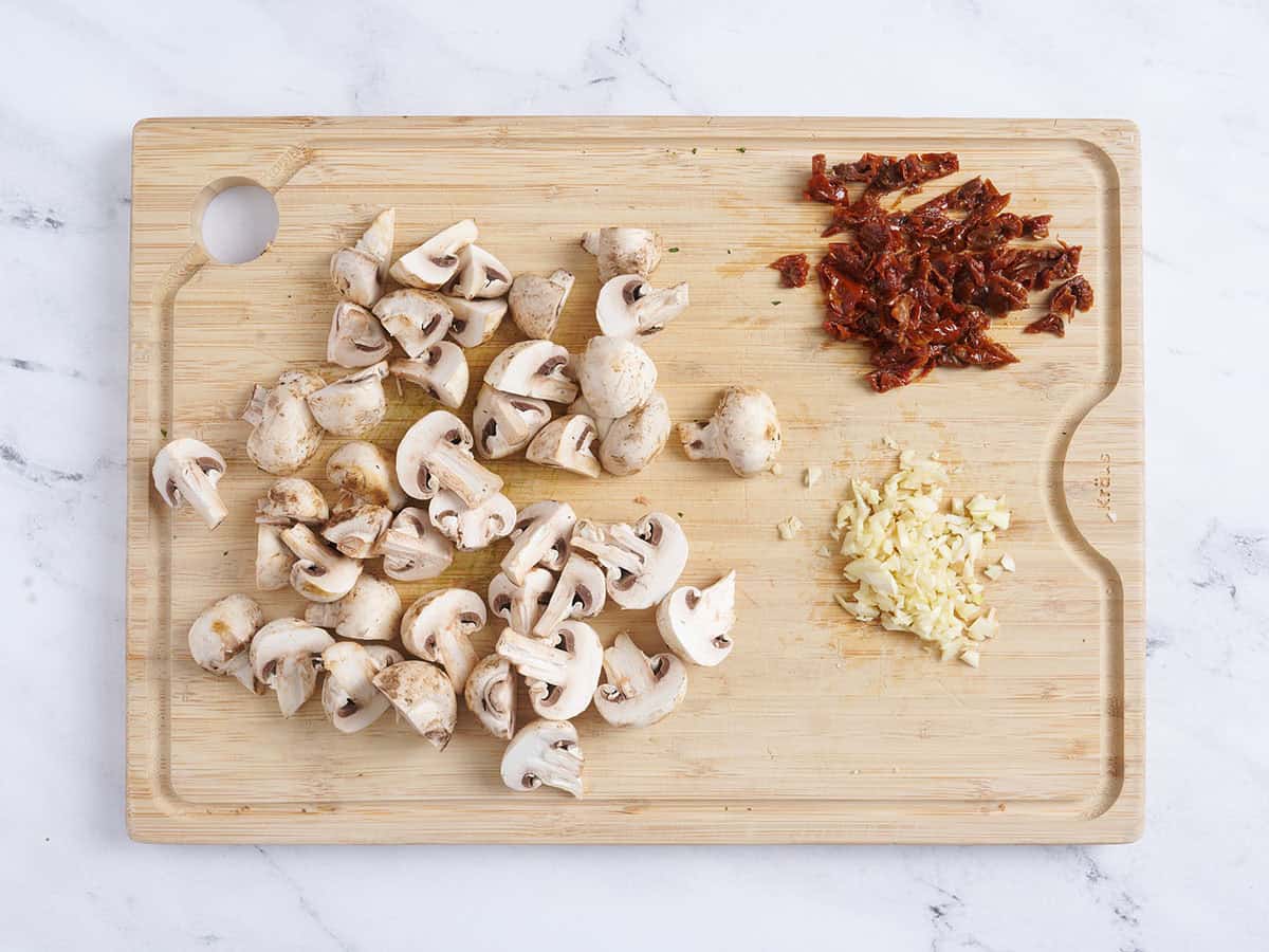 Sliced mushrooms, sundried tomatoes, and garlic on a wooden cutting board.