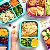 Overhead view of a selection of packed bento lunchboxes with a childs hand taking some cheese.