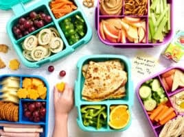 Overhead view of a selection of packed bento lunchboxes with a childs hand taking some cheese.