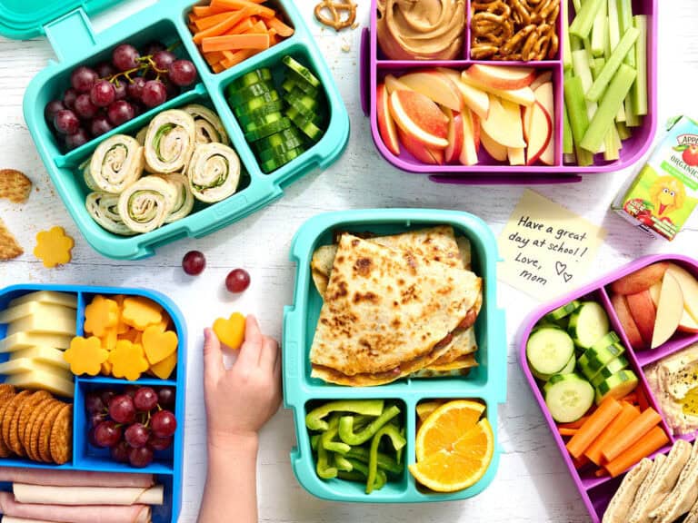 Overhead view of a selection of packed bento lunchboxes with a childs hand taking some cheese.