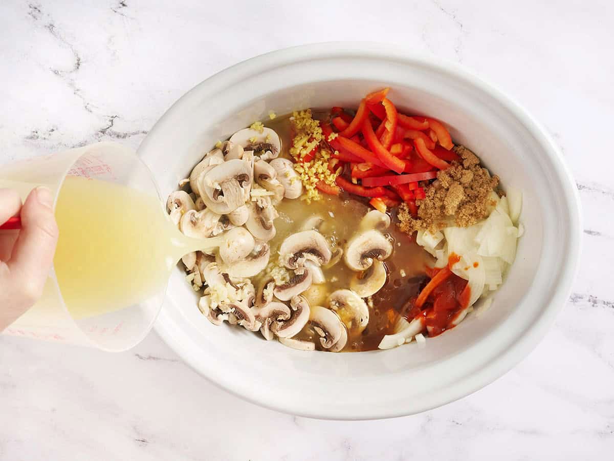 Sliced mushrooms, peppers, onion, brown sugar, and soy sauce in a slow cooker with broth being poured in.