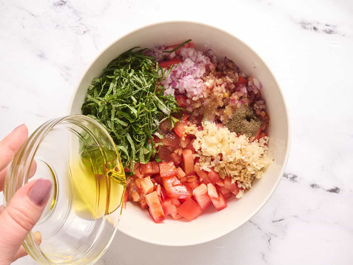 Diced tomatoes, sliced fresh basil, diced red onion, seasonings, minced garlic in a bowl with olive oil being poured in.