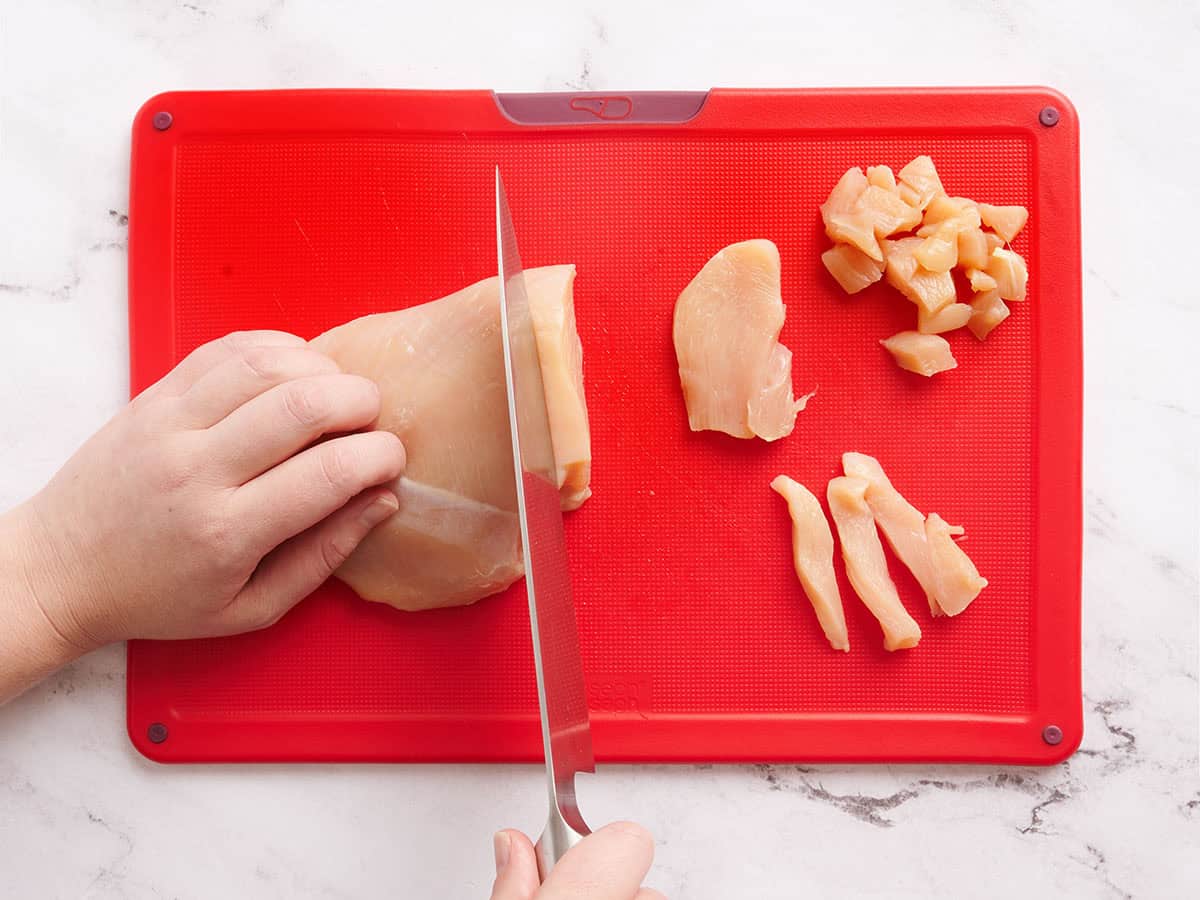 A hand using a knife to cut chicken breast into chunks.