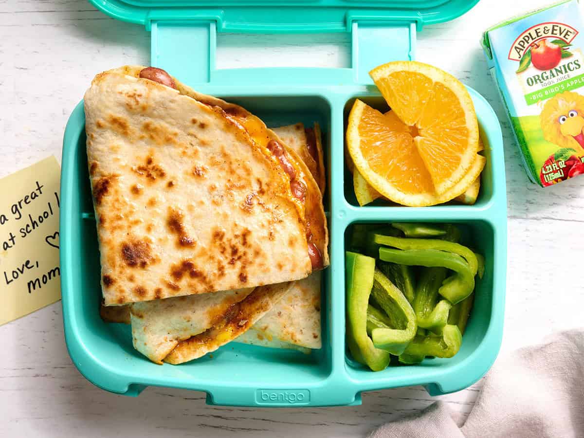 An overhead view of cheesy kidney bean quesadillas in a lunch box with orange slices and green bell pepper slices.