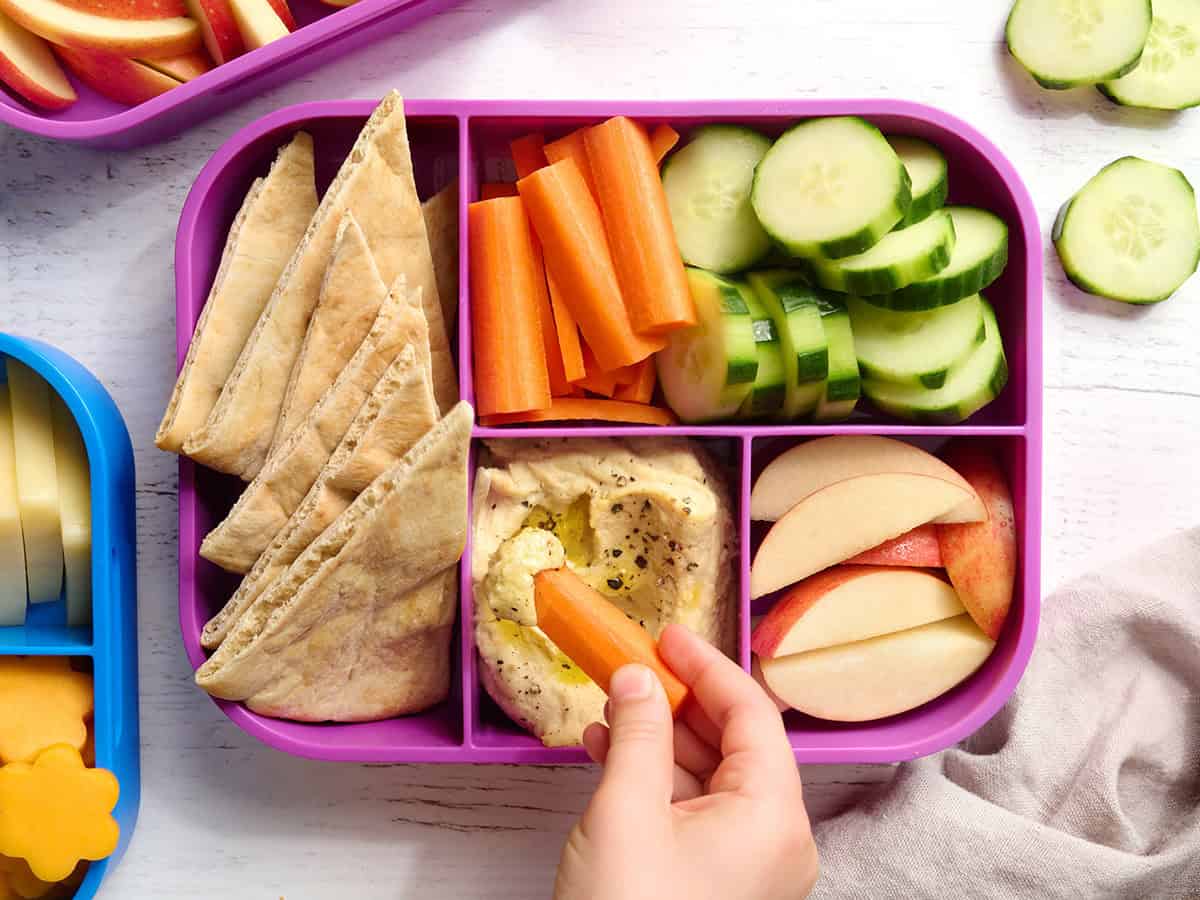 Overhead of a hummus lunch box with hummus, carrot sticks, pita, cucumber, and apples, with a childs hand dipping carrot sticks into hummus.