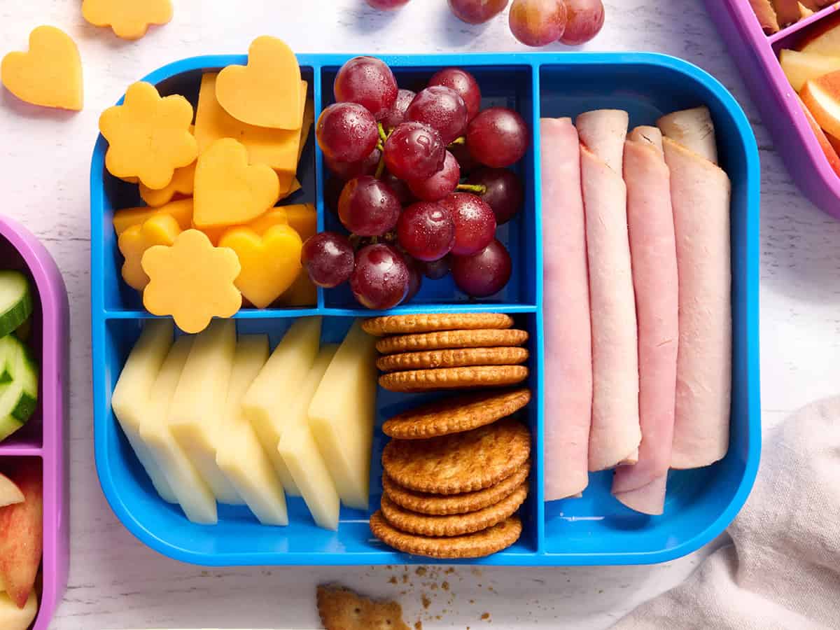 An overhead shot of a cheese board with crackers, ham, cheese and grapes, lunch box.