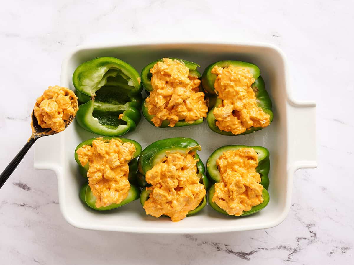 Green bell pepper halves in a baking dish with buffalo chicken filling being added.