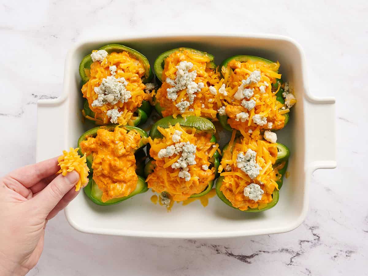 Green bell pepper halves stuffed with buffalo chicken filling, with a hand sprinkling cheddar cheese and blue cheese on top.