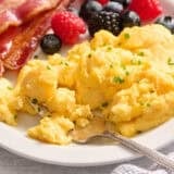 Overhead side view of cottage cheese eggs on a plate with bacon and a fruit salad.