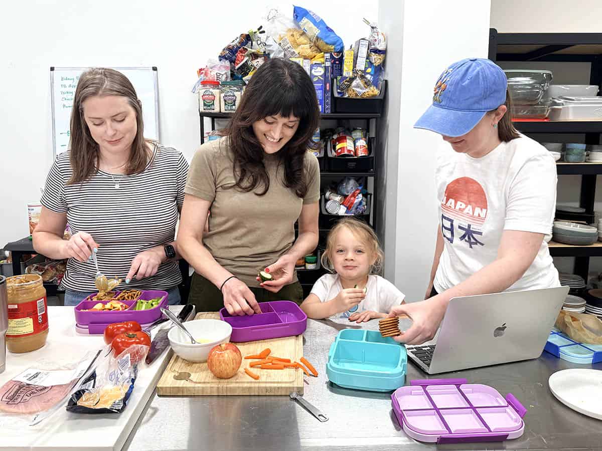 Three women and a little girl stood at a workbench packing lunch boxes.
