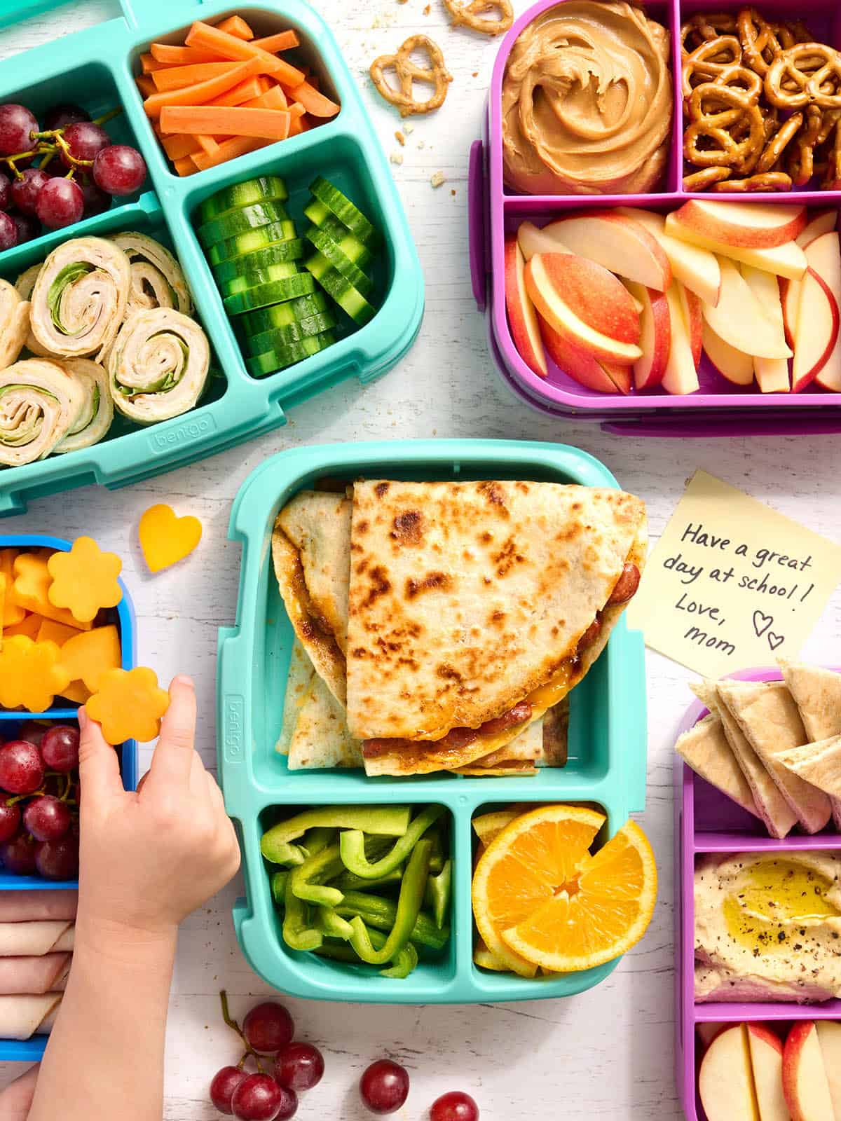 Overhead view of a selection of packed bento lunchboxes with a childs hand taking some cheese.