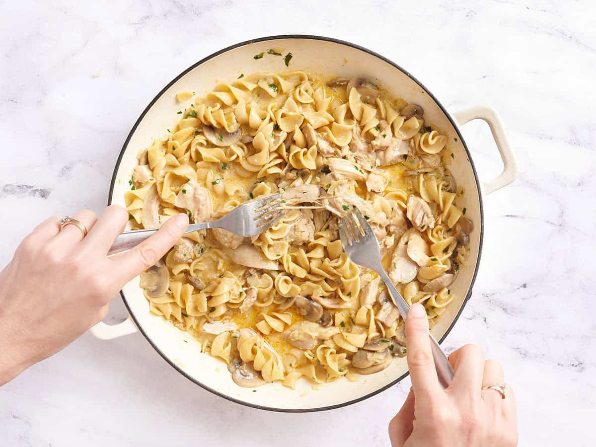 Chicken thighs being shredded into chicken stroganoff in a skillet.