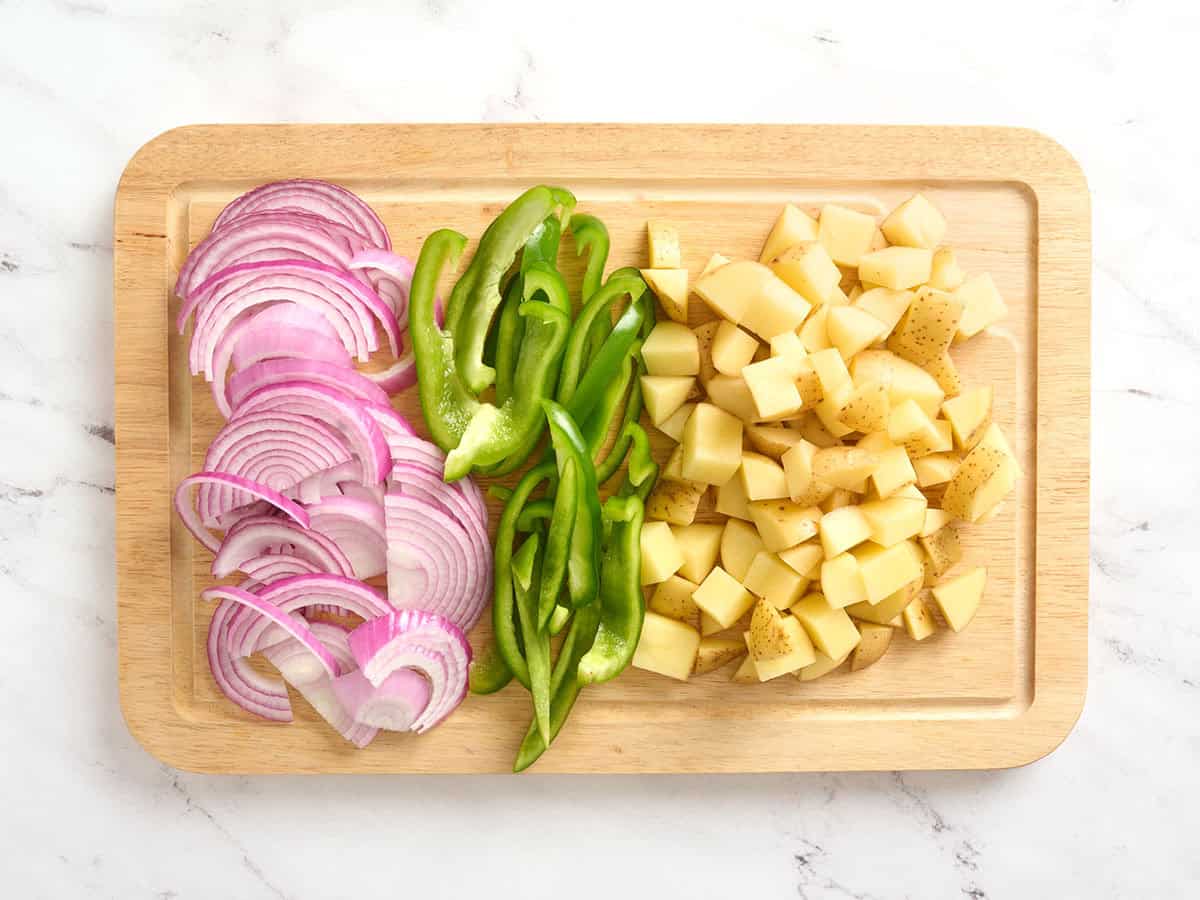 Sliced red onions, green bell peppers and diced potatoes on a cutting board.