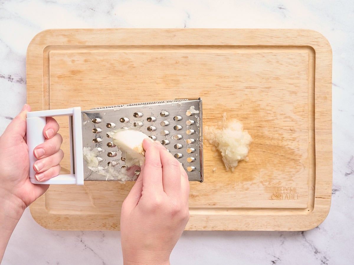 Hands using a cheese grater to grate a white onion.